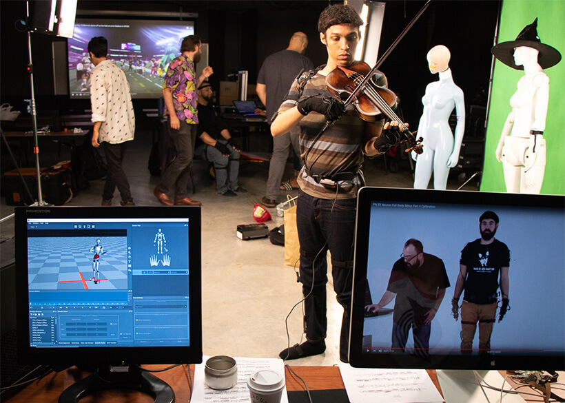 Student playing violin in a production studio in front of a green screen and wearing motion capture equipment so you can see a 3D model of the student on a computer screen in front
