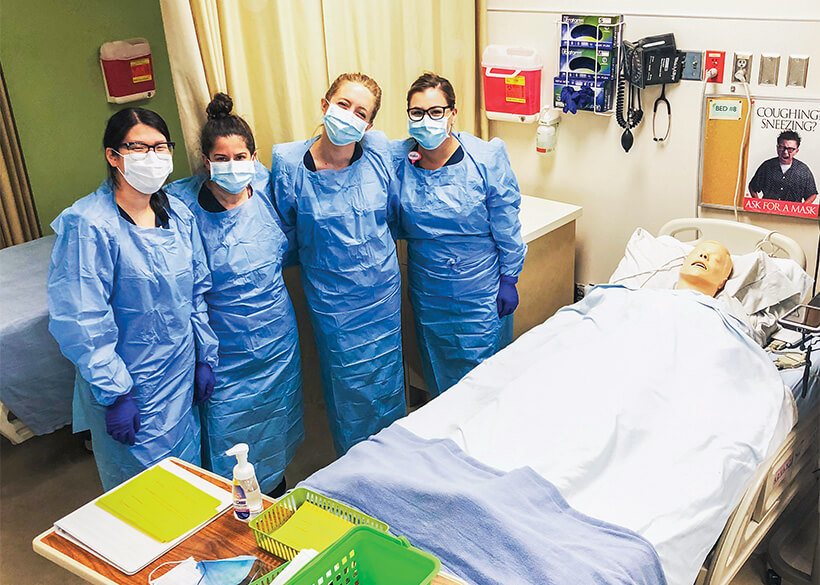 Four students in scrubs in a medical setting with a practice dummy patient on the exam table